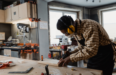 A person is a professional woodworking shop using a router on a wooden board. They are wearing a work apron, safety googles, a mask and ear protection.