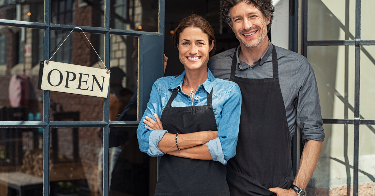 Two people wearing aprons outside a business storefront with an Open sign, smiling.