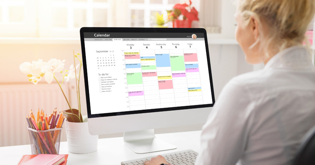 A woman working on a digital planner on her computer monitor. To the left of the monitor is a cup of pencils.