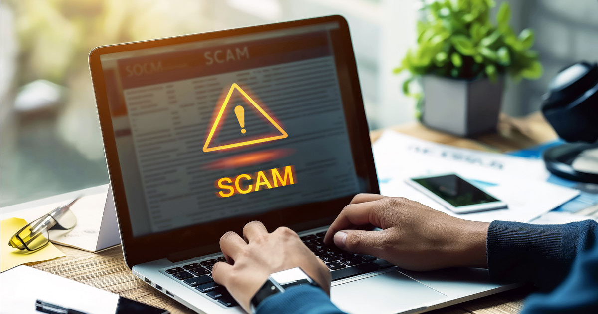 A desk with someone typing on a computer. The computer shows a screen with a triangle with an exclamation mark with scam written below.