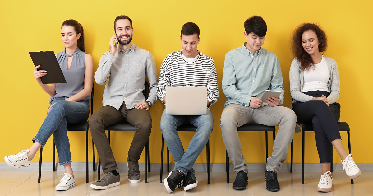 Five people sitting in a row of chairs, waiting for a job interview. One person is working on a laptop, one is looking at a notepad, one is talking on a phone, and one is looking at a clipboard.