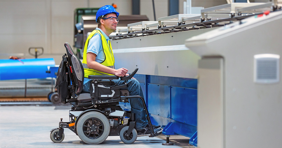 A person using a wheelchair at a manufacturing job. He is wearing a hard hat and safety vest.