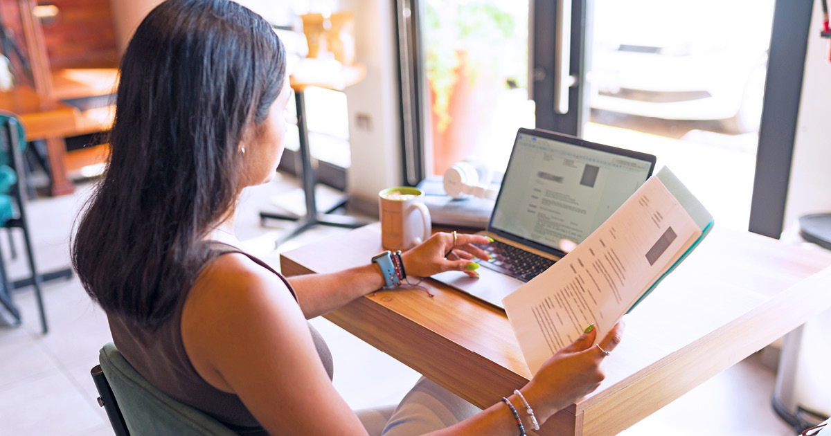 A person working on a laptop and holding sheets of paper in her right hand.