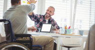 A person who is a wheelchair user high fives another person across a desk.